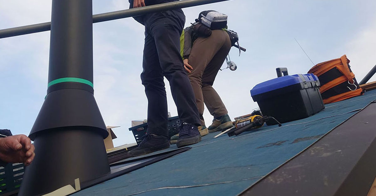 屋根の上に立つ職人の足元と、取り付けを終えた煙突トップ、屋根に置かれた工具箱