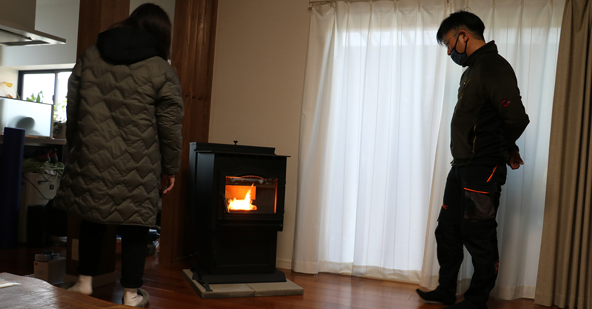 リビングに設置され火が入ったペレットストーブと、そばで暖まる住まい手、立ち会う施工担当者